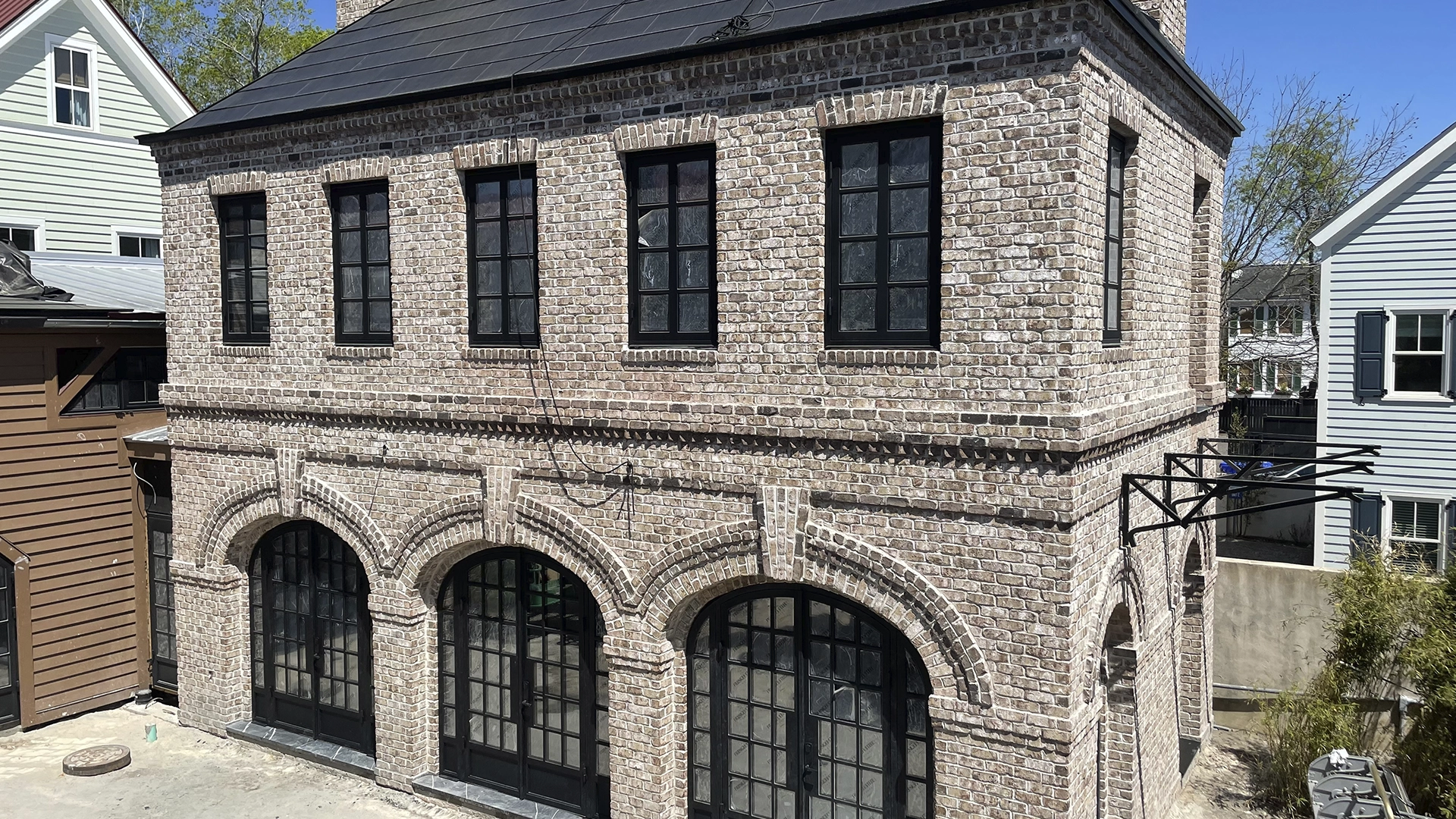 A two-story brick building with black-framed arched windows and a sloped roof, flanked by neighboring houses.